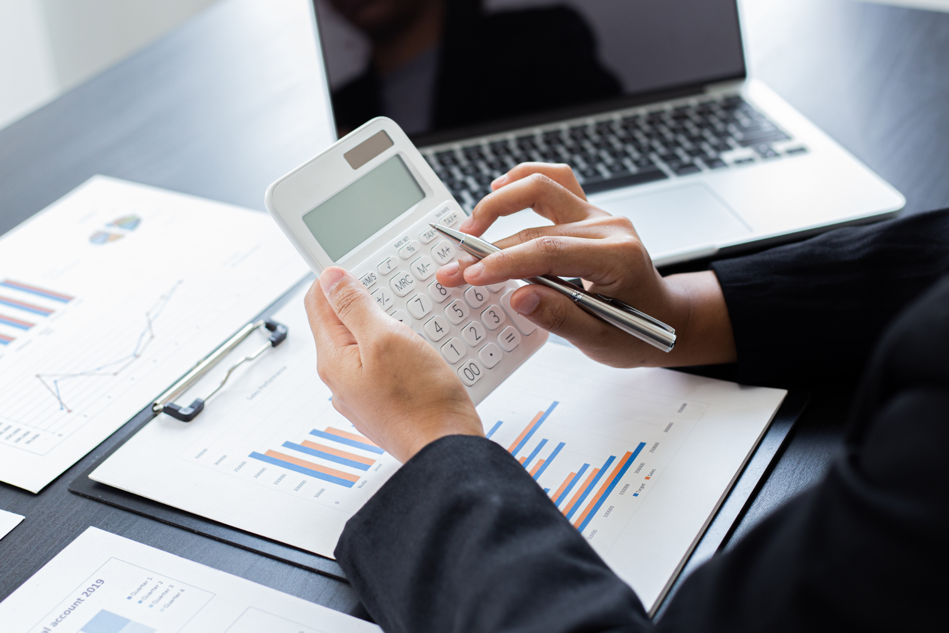 Hands using a calculator over financial charts beside a laptop on a desk