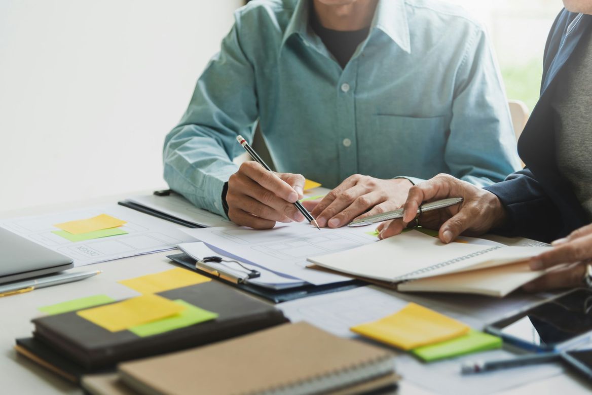 Two people review paperwork at a desk with notebooks, sticky notes, and a laptop.