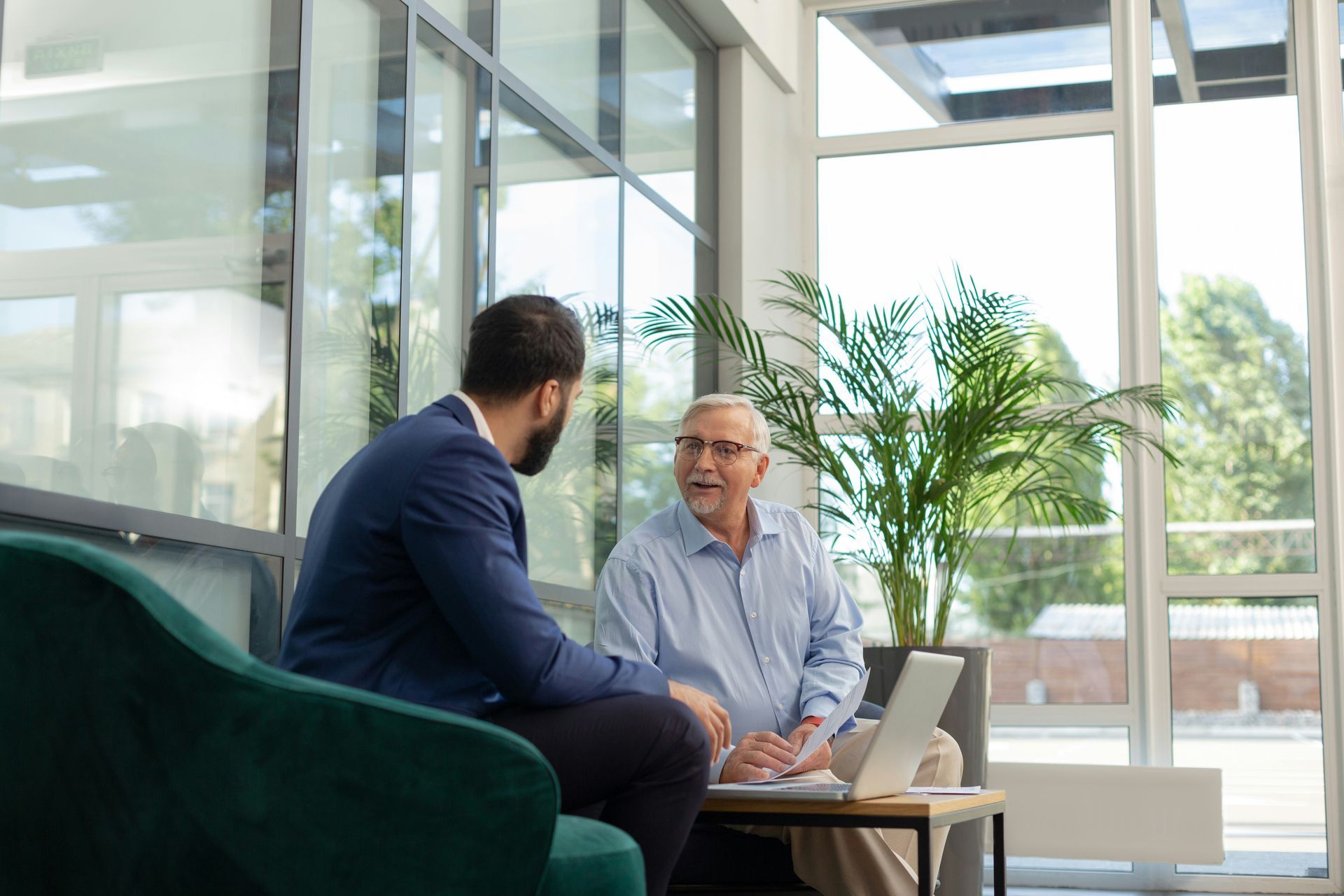 Two people talking in a bright office lounge beside a laptop and large windows.