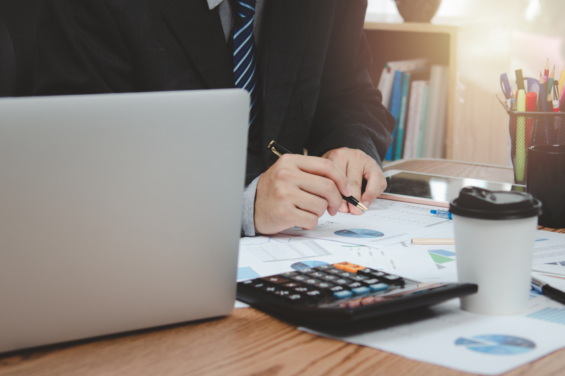 Person in business attire reviewing charts at a desk with a laptop, calculator, and coffee cup
