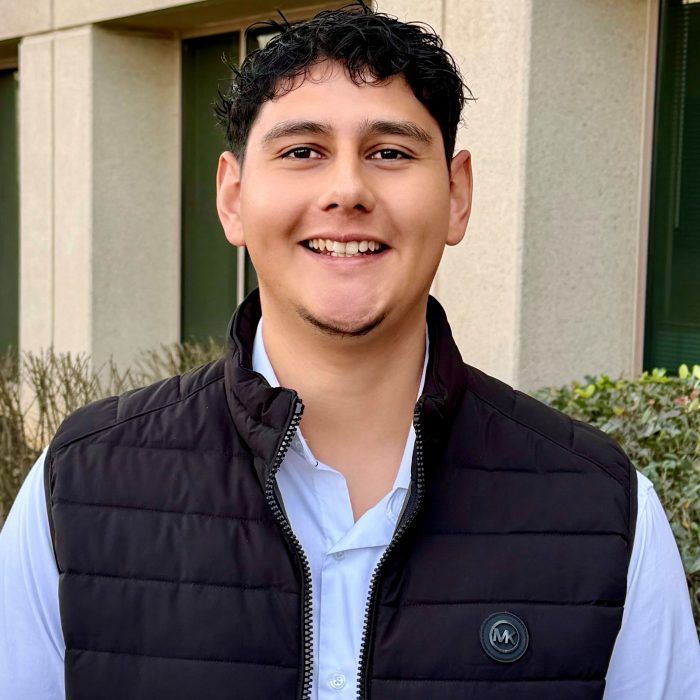 Smiling person in a black vest and white shirt standing outdoors by a building.