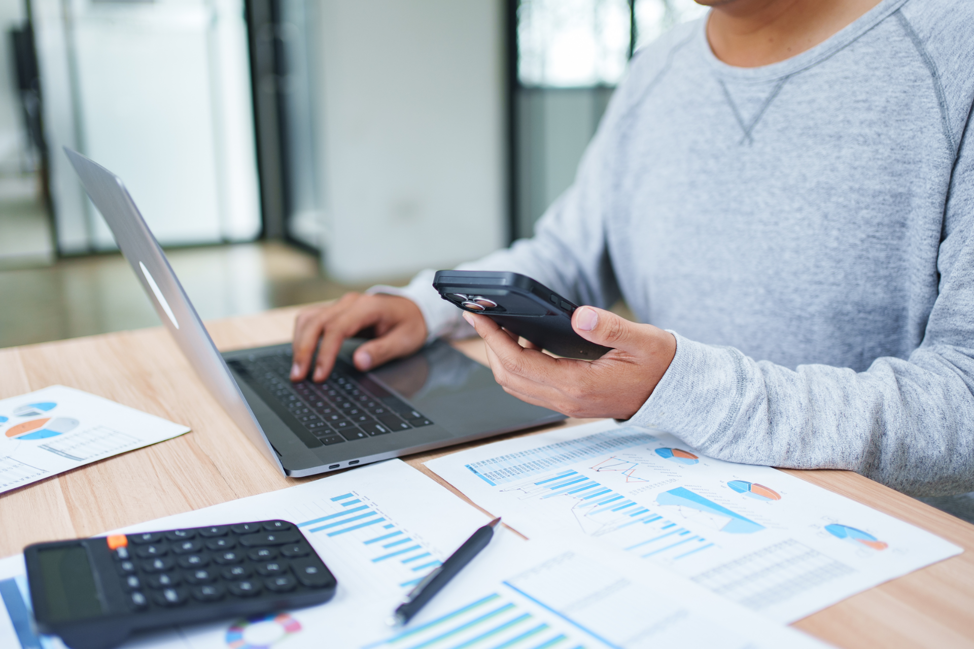 Person using a laptop and smartphone at a desk with financial charts and a calculator.