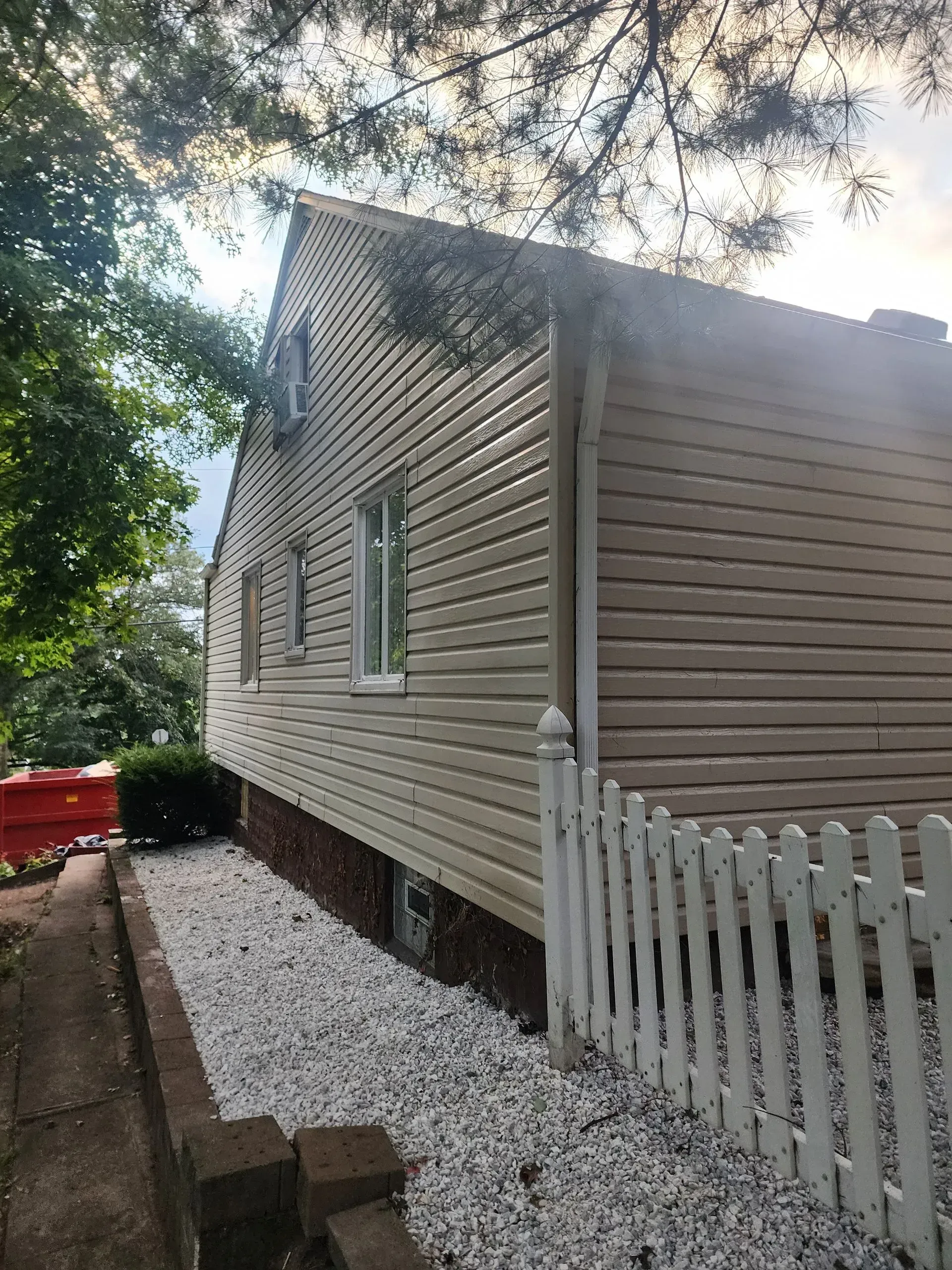 Side view of a beige house with white trim, gravel path, and white picket fence under trees.