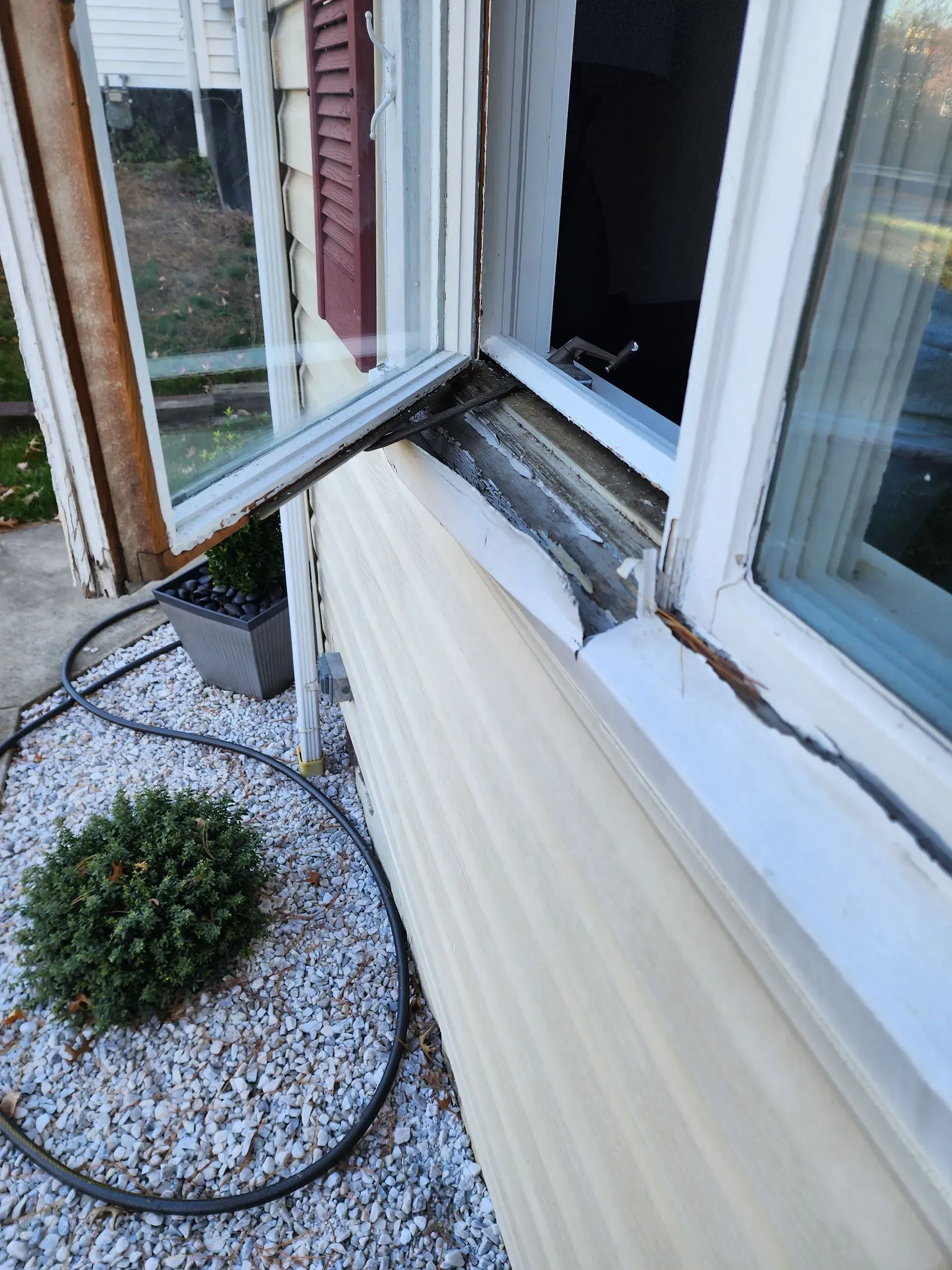 Open window with damaged sill above a gravel garden bed and black hose