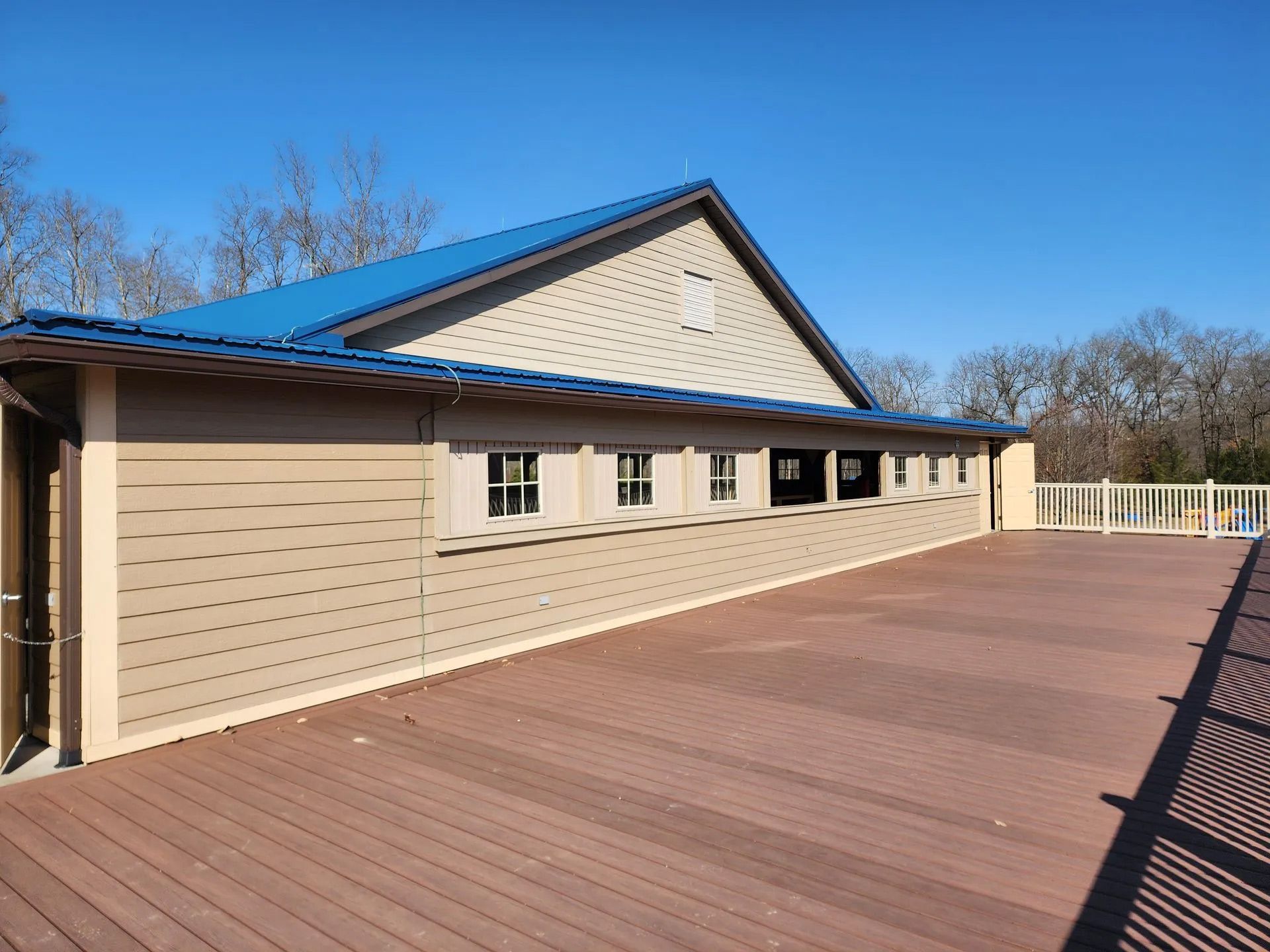 Beige building with blue roof beside a wide wooden deck under a clear blue sky