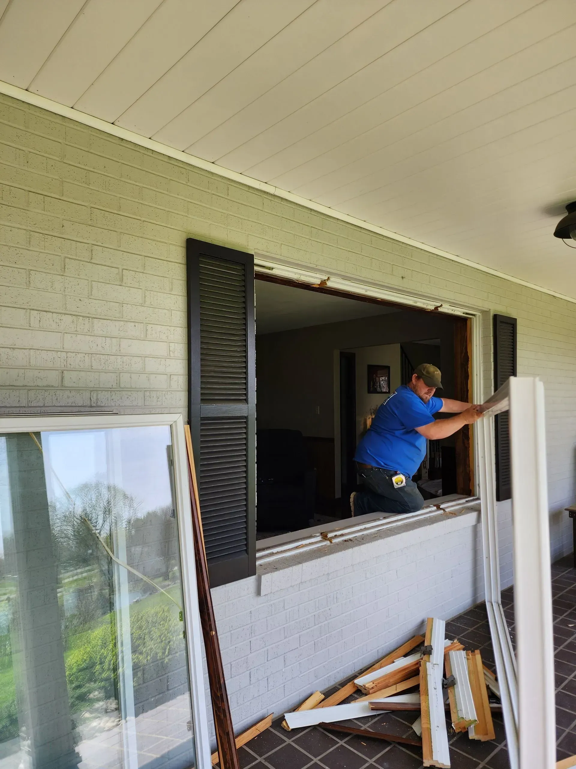 Person leaning out of a house window, working on the frame beside stacked wood and tools on the porch.