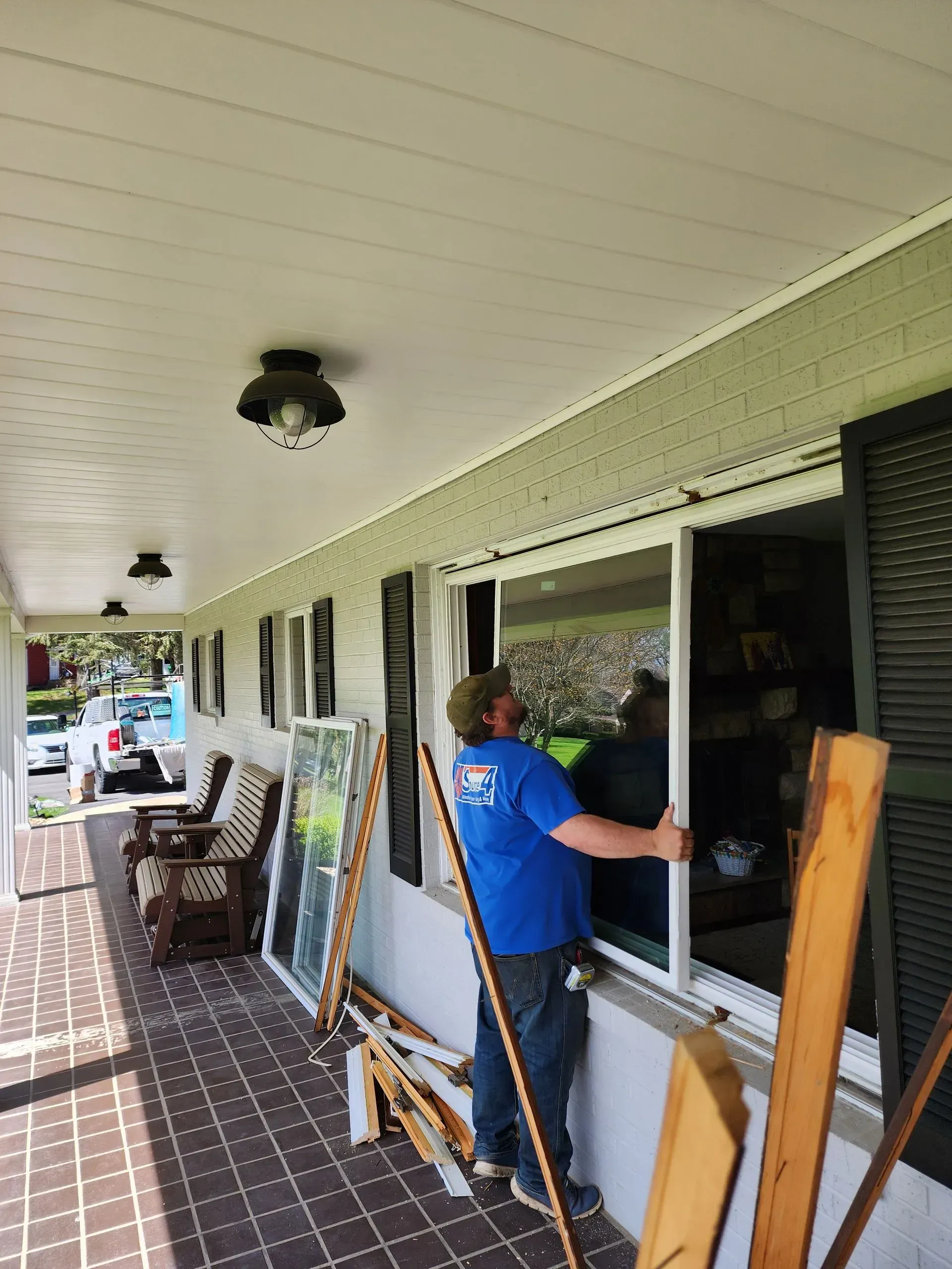 Person in blue shirt installing a window on a covered porch with tools and stacked glass panes.