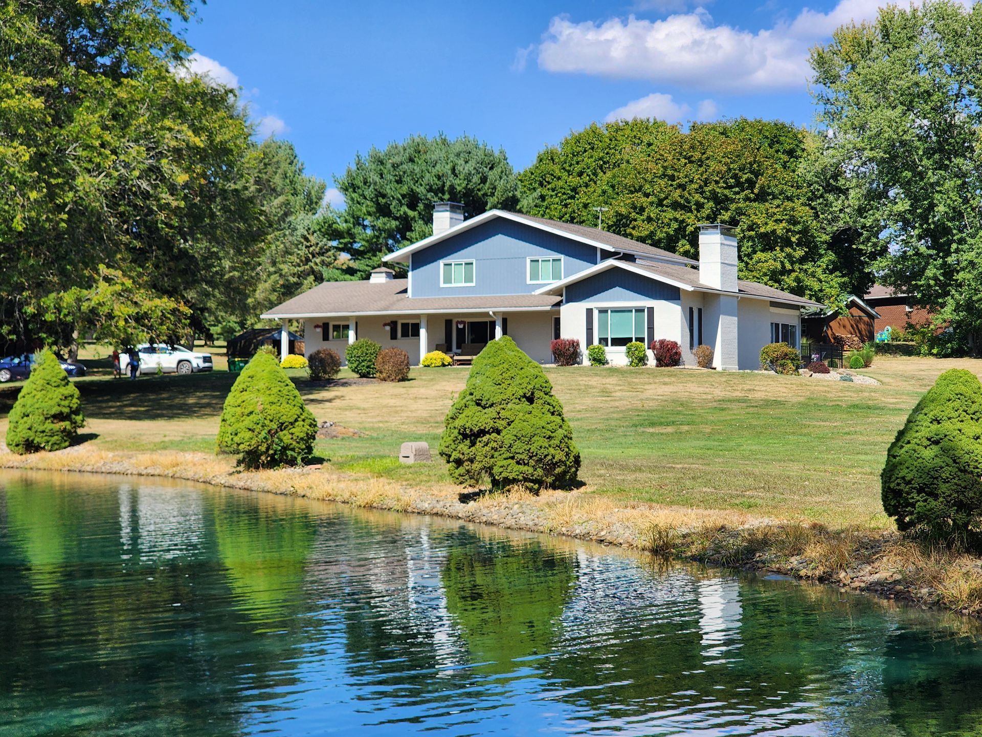 Lakeside house with green lawn, trees, and reflections in calm water under a blue sky.