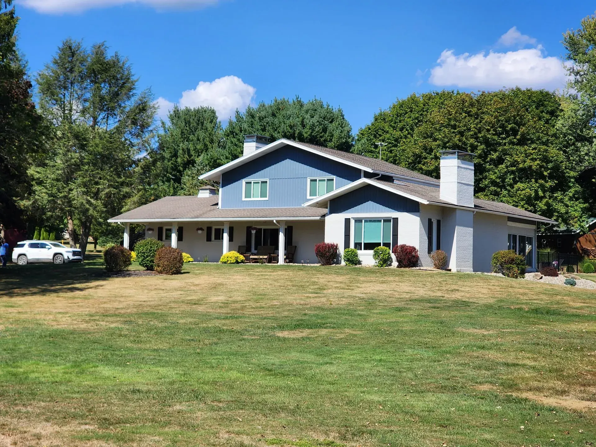 Large gray house with white trim on a grassy lawn under a blue sky, surrounded by trees