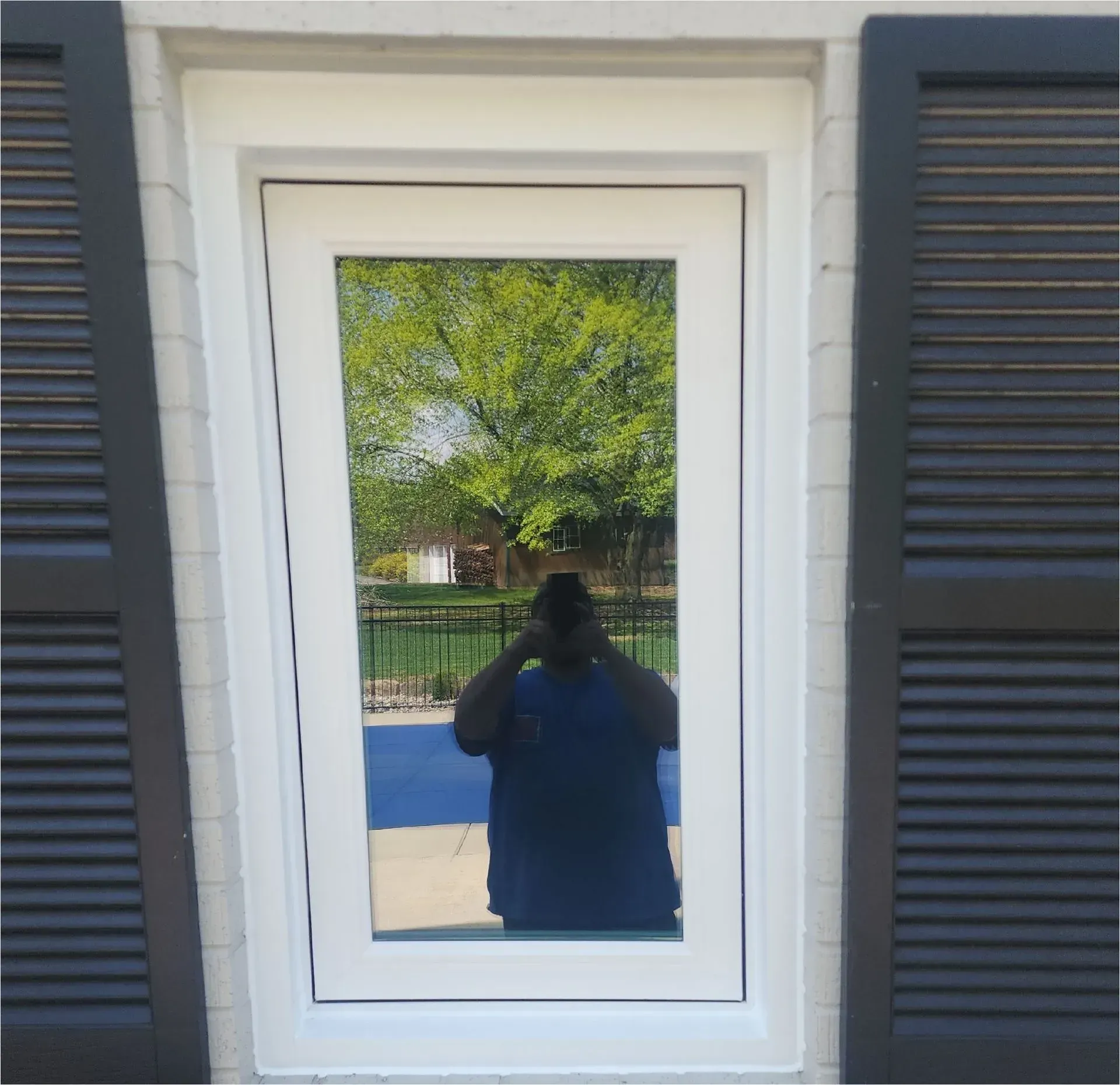 Person reflected in a white-framed window, standing before a building with dark shutters and trees behind.
