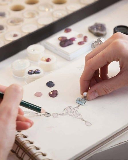 Hands arranging small blue stones over a sketch on a white worktable with art supplies nearby