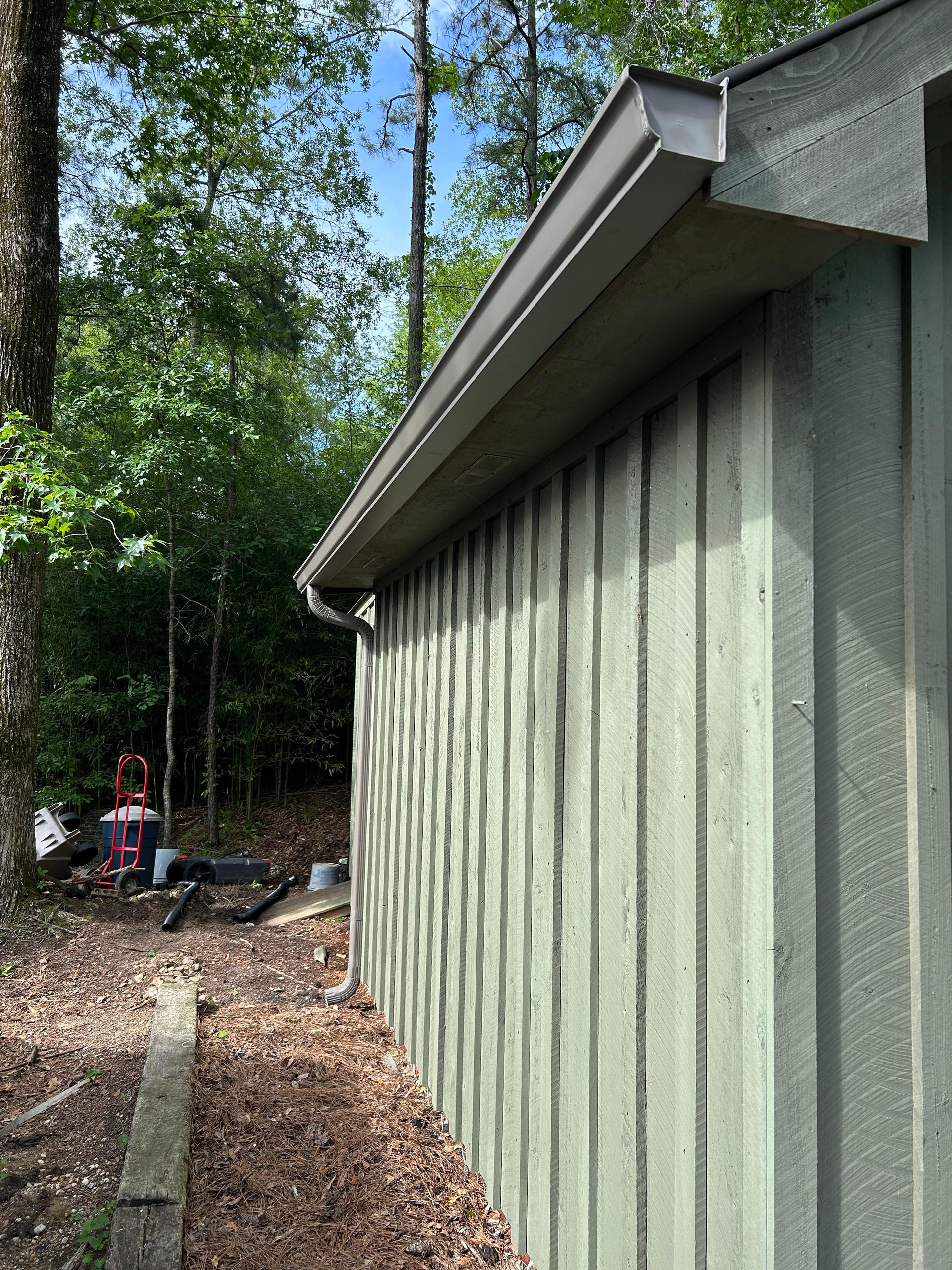Gray house siding and roof eave beside a wooded yard, with a dirt path and leaves below.