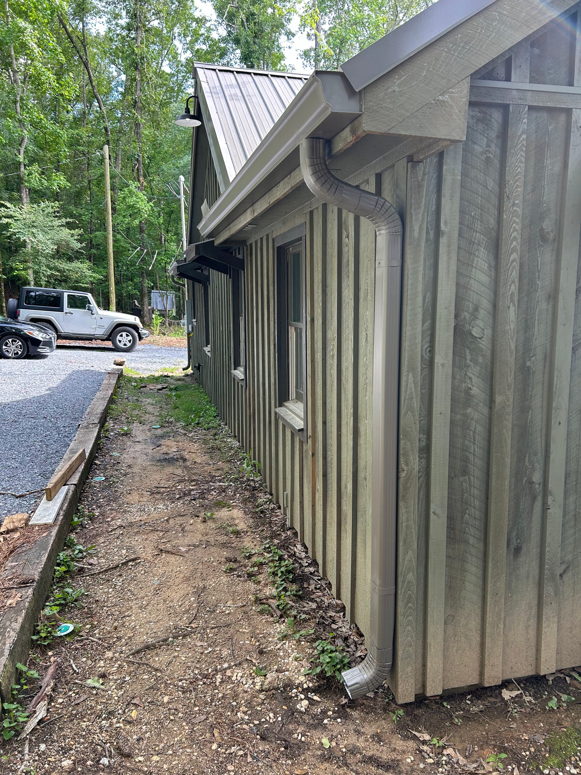 Side view of a rustic wooden building with metal roof, windows, and downspout beside a gravel driveway in a wooded area