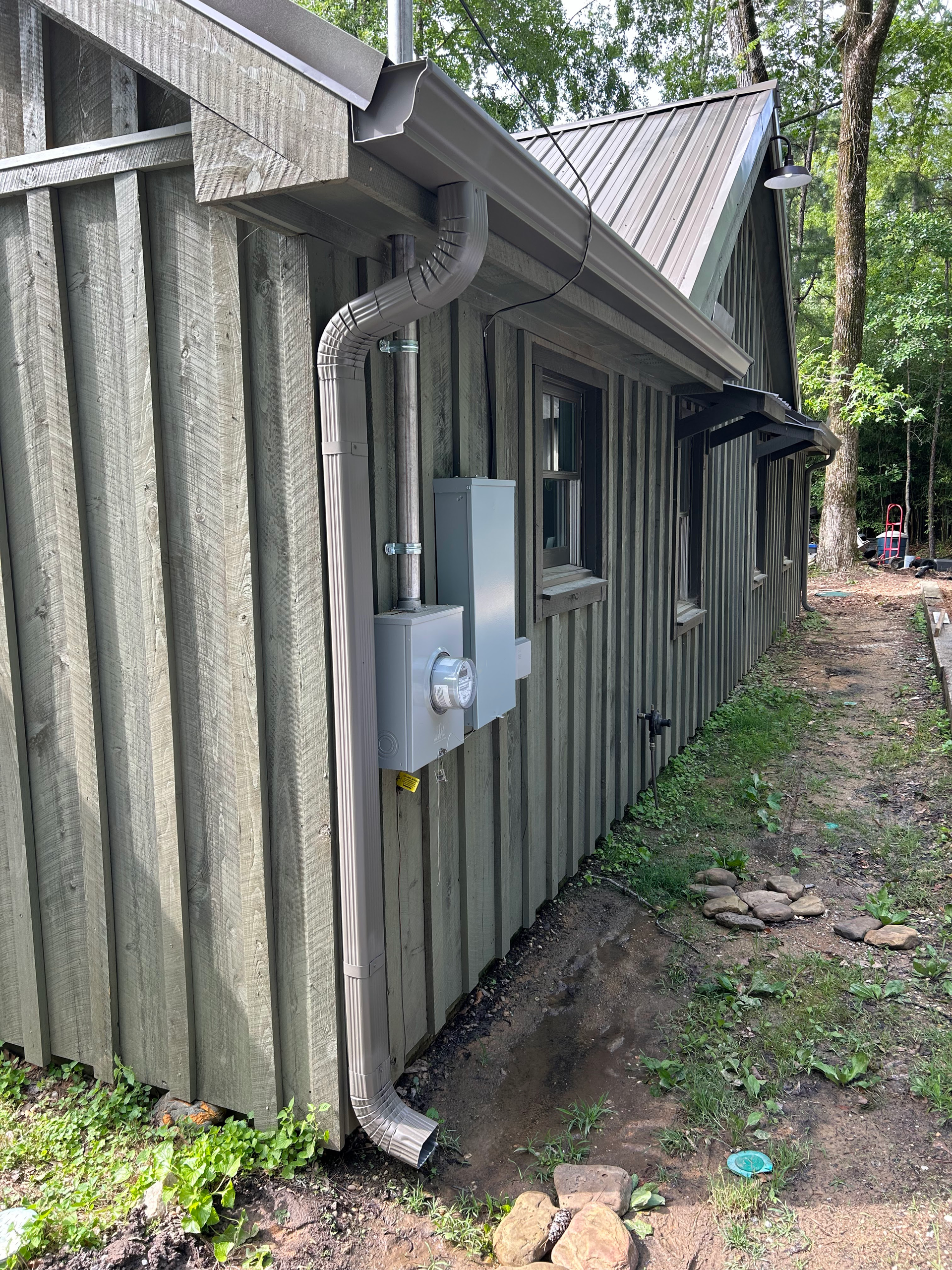 Weathered wooden building with an electrical meter and conduit along a narrow dirt path beside trees.