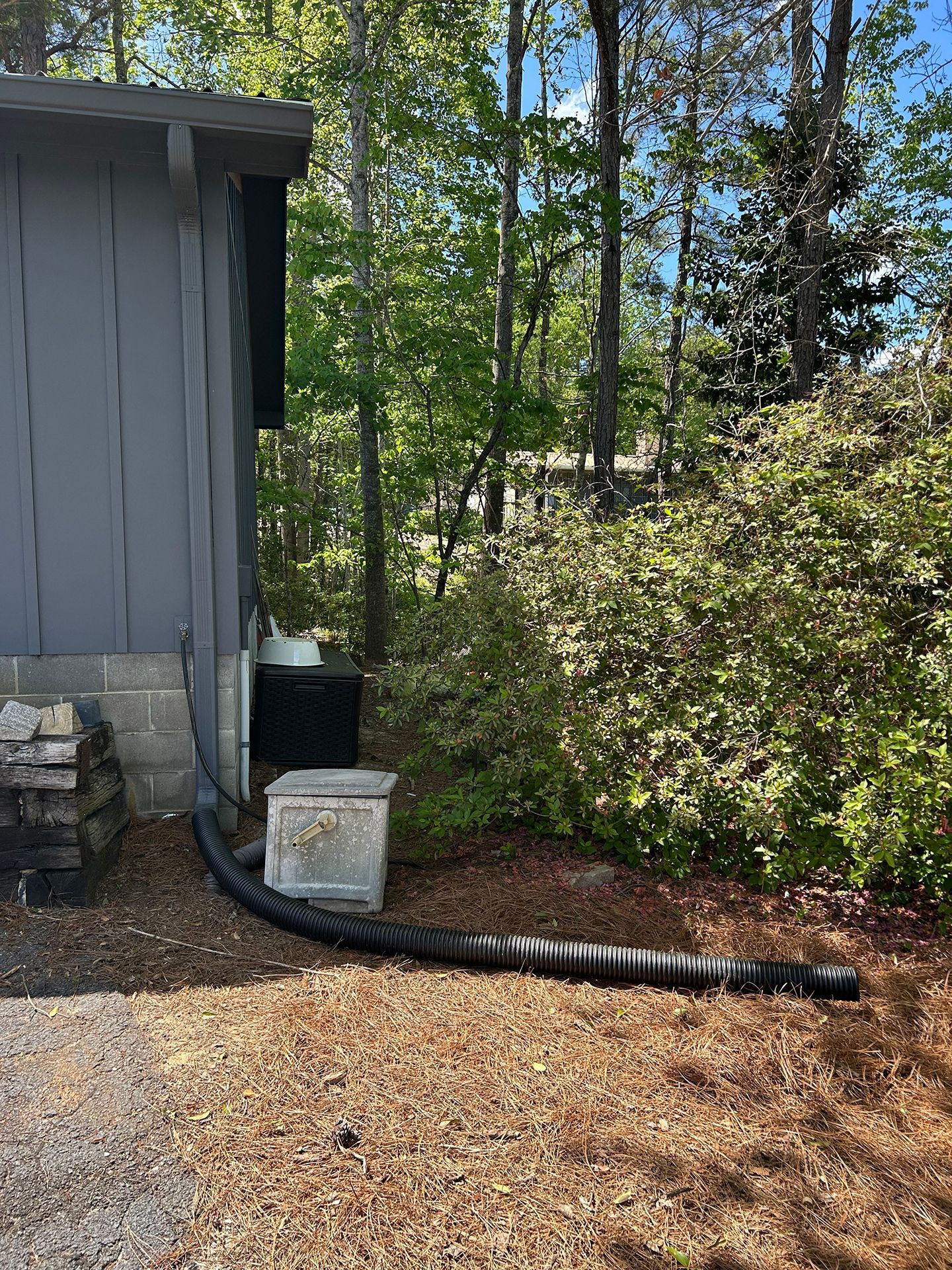 Side of a gray house with a black drainage hose, utility boxes, and trees in a wooded yard.