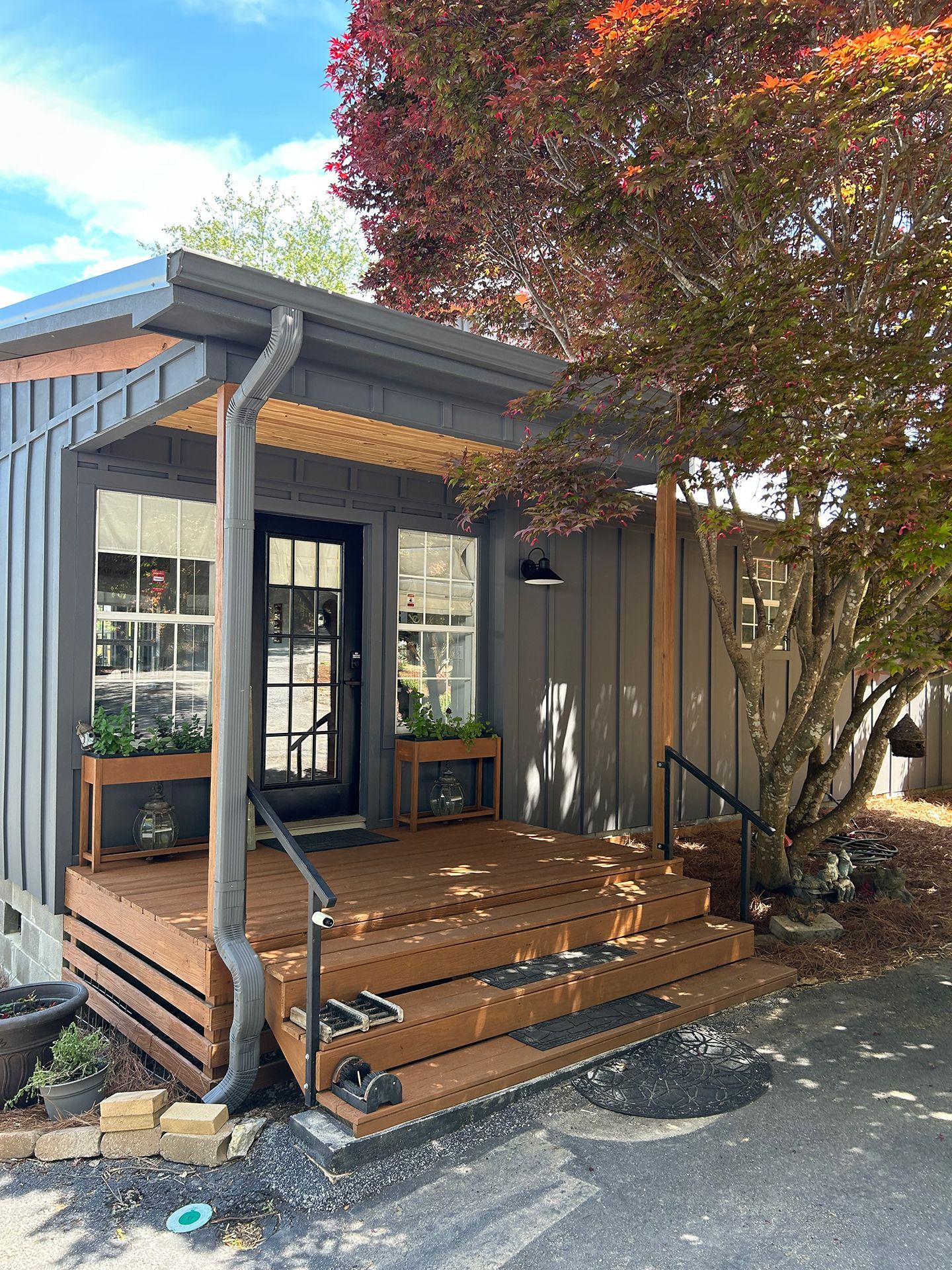 Small front porch with wooden steps, railings, and a tree beside a gray house under a blue sky.