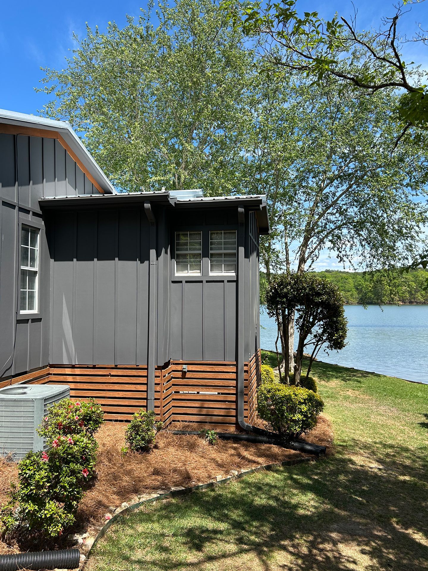 Gray lakeside cabin with black trim, tree overhead, and a small garden by the water