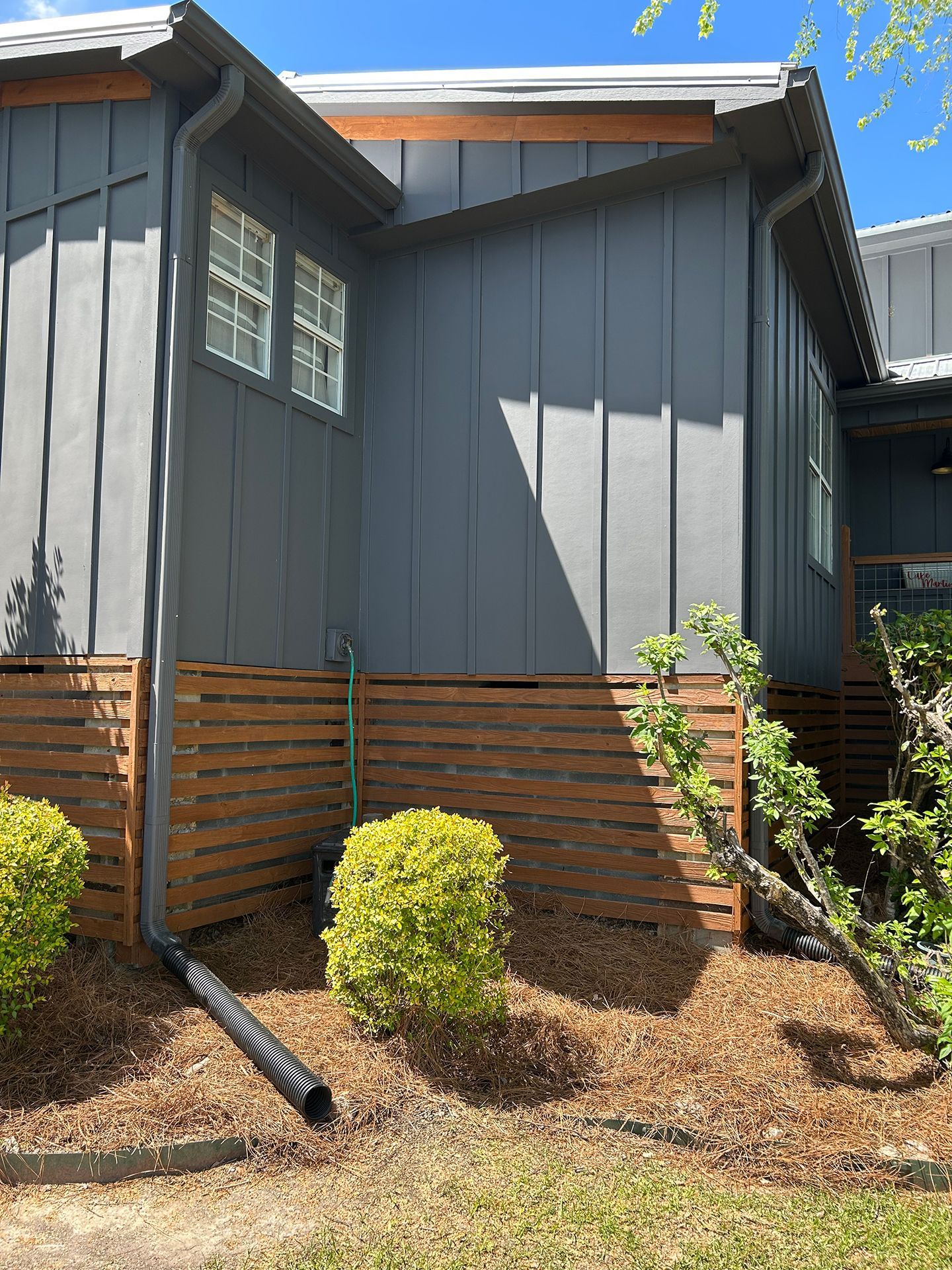 Gray house exterior with horizontal wood fence, small shrubs, and mulched yard in bright sunlight