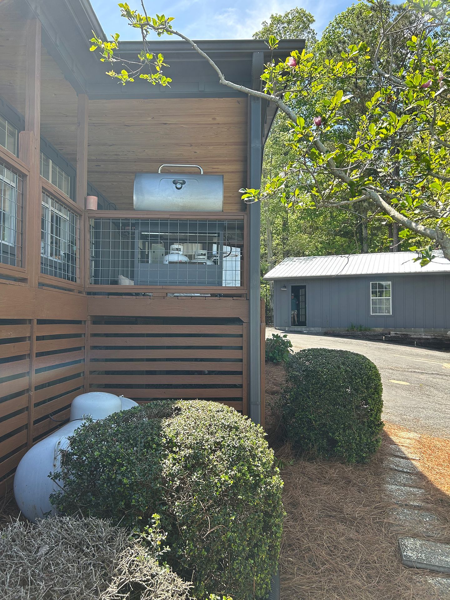 Wooden apartment balcony with railings, shrubs, and a gravel path beside a small outbuilding