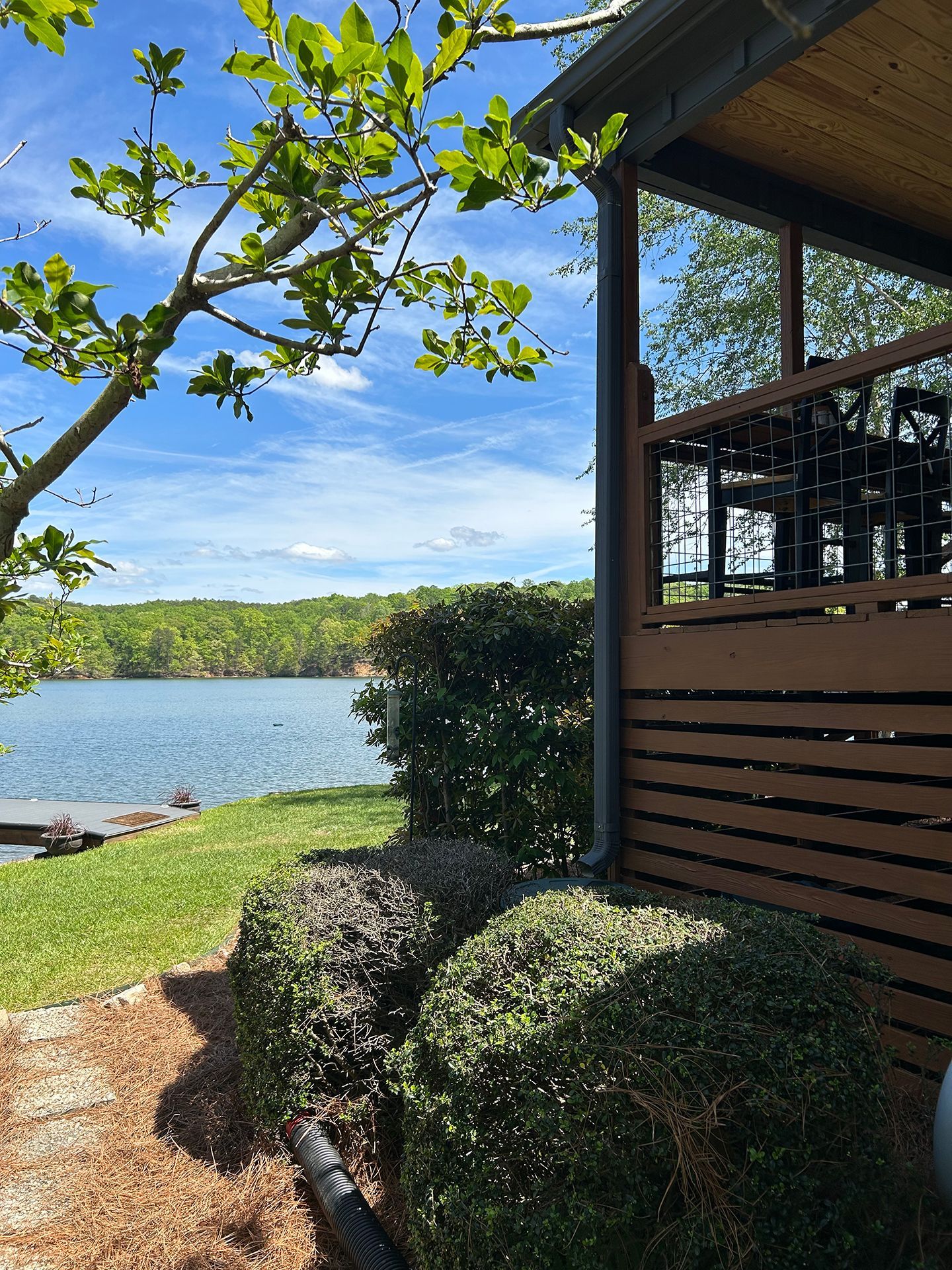 Lake view from a shaded patio with trimmed hedges and a wooden deck under a blue sky.