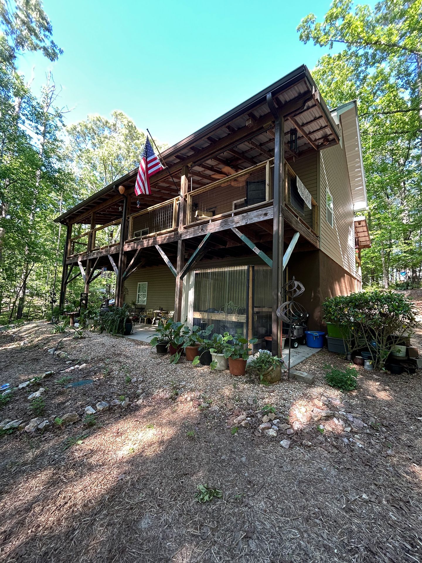Rustic elevated cabin with a wraparound porch, American flag, and trees surrounding it