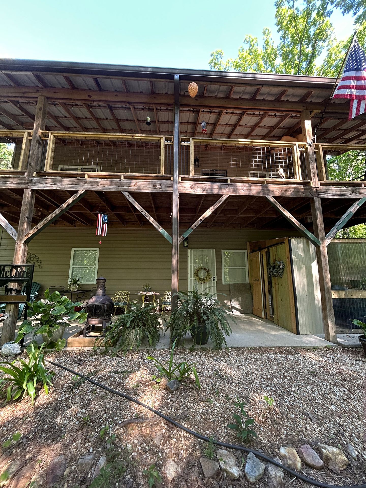 Rustic cabin porch with wooden beams, green siding, gravel path, and potted plants under a metal roof.