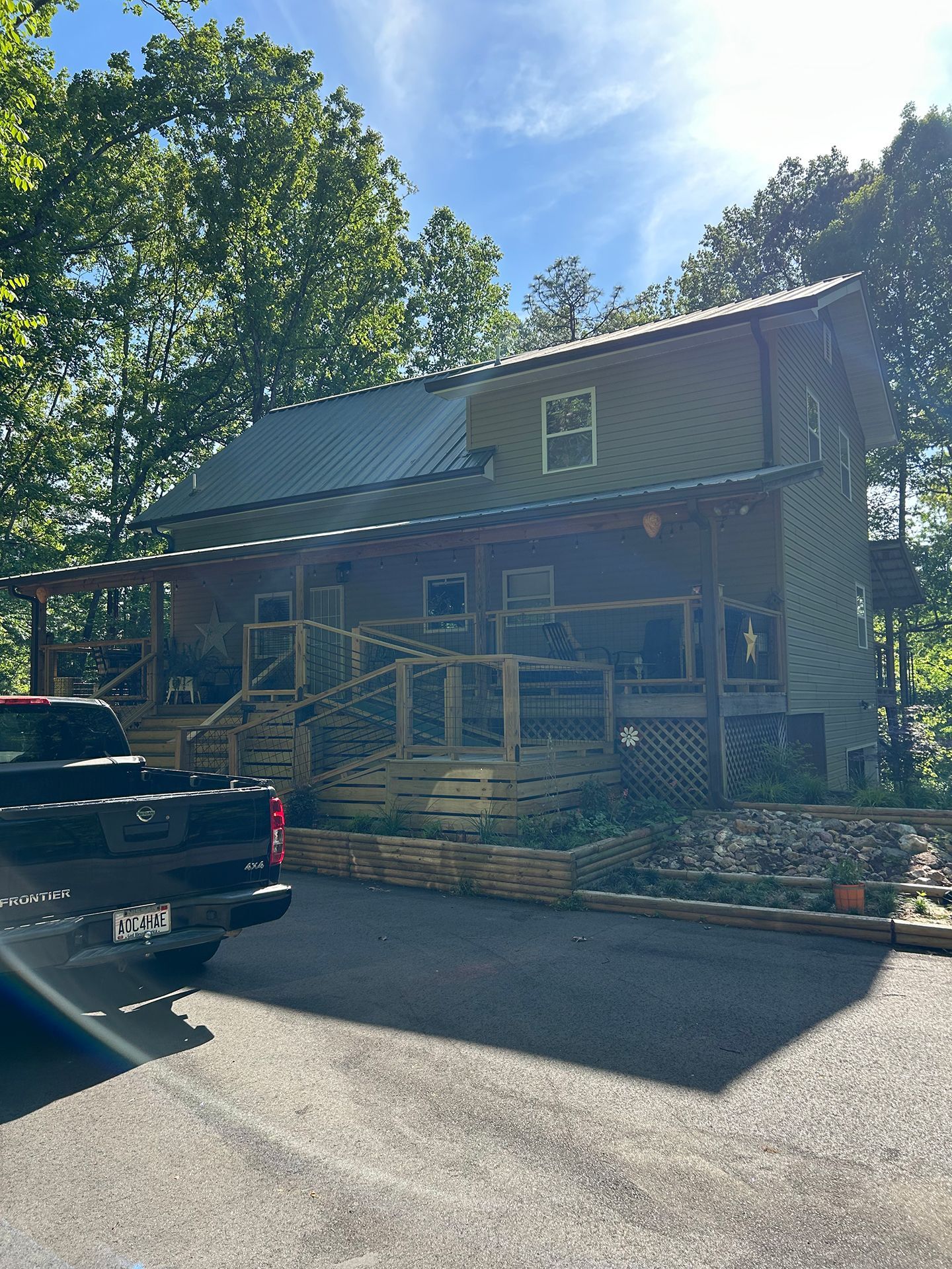 Two-story house with front porch and stairs, seen from a driveway beside a black car in bright sunlight