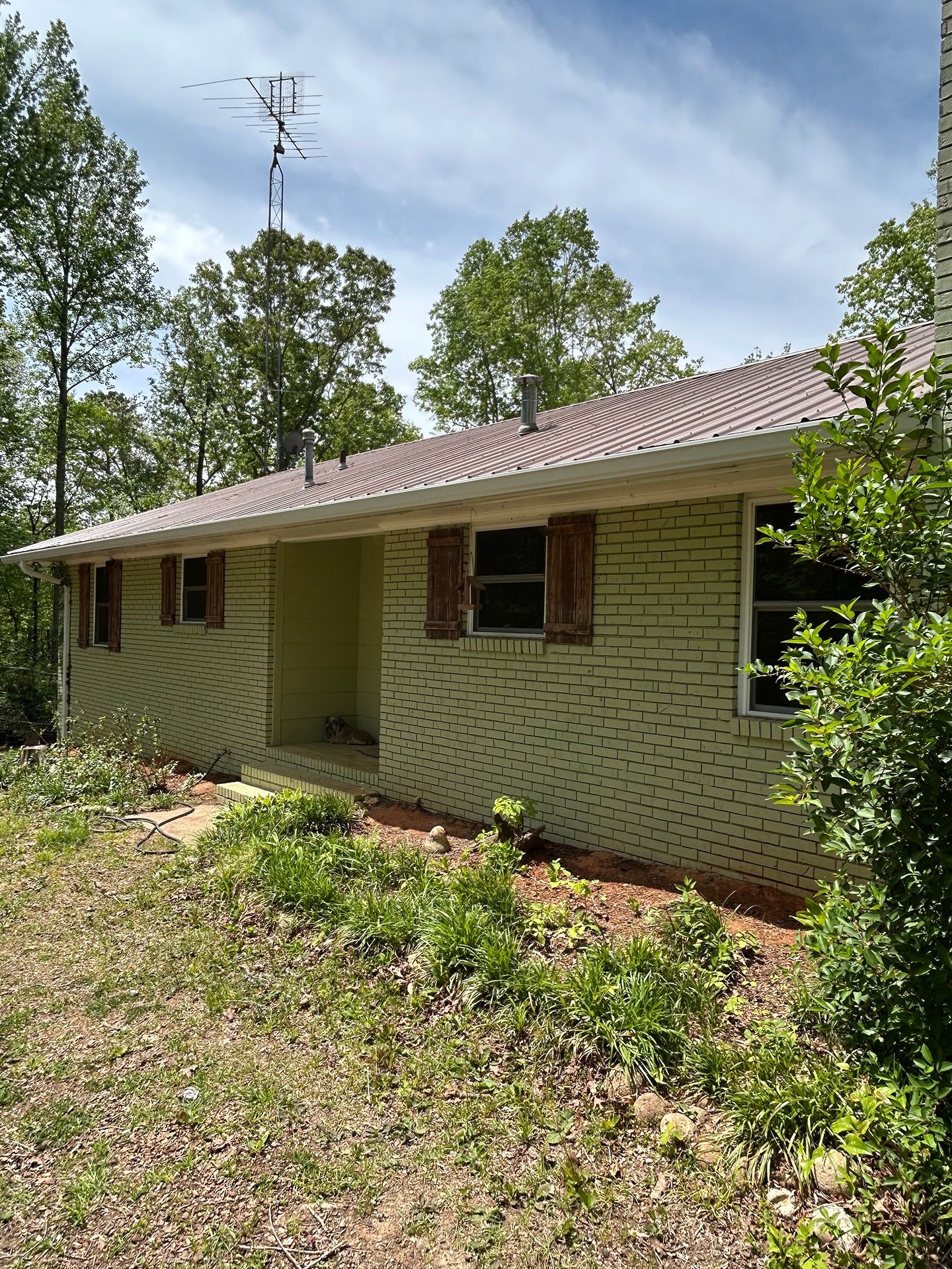 Single-story white brick house with red roof, front porch, and overgrown yard in a wooded setting