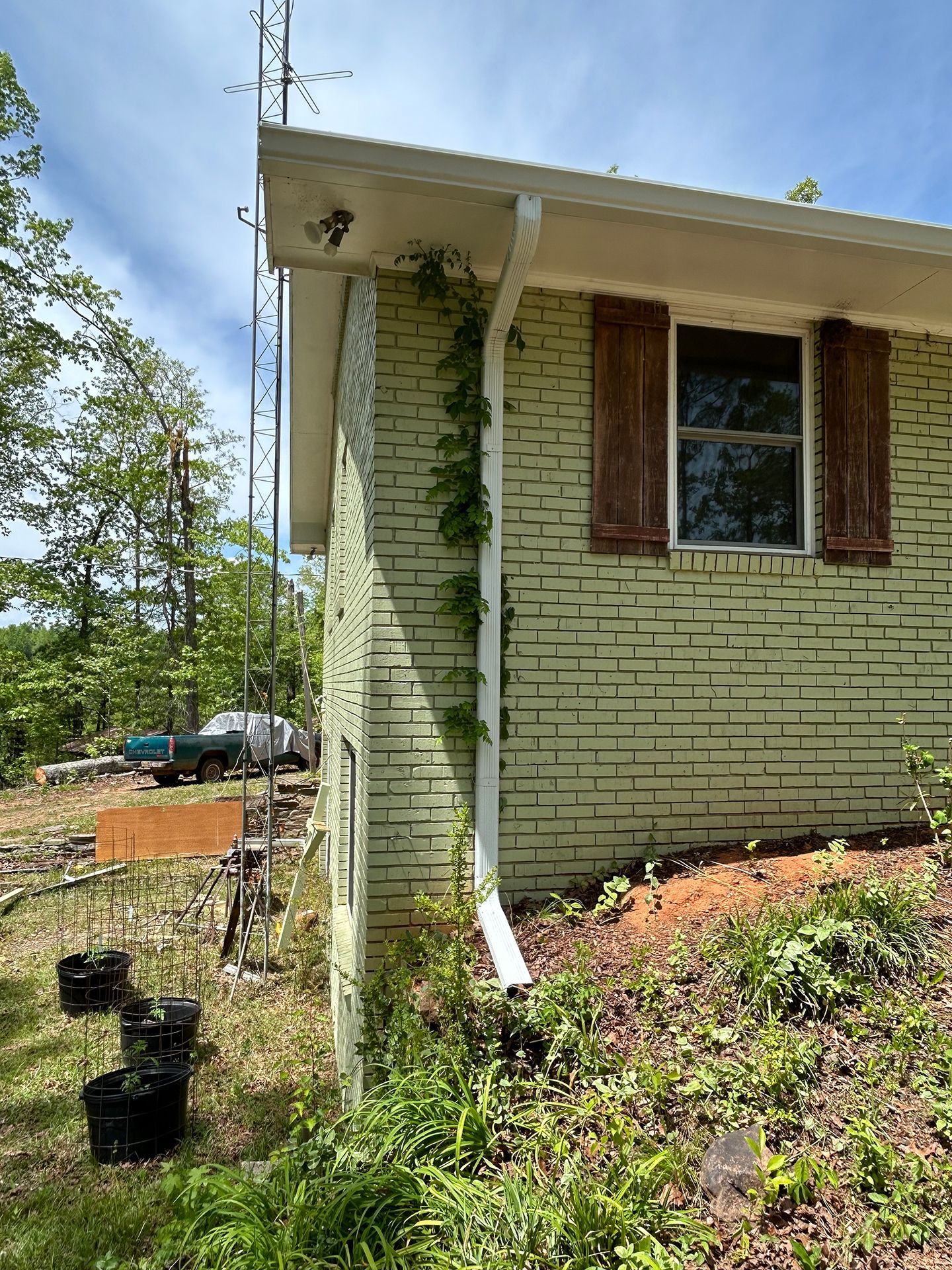 Side view of a beige brick house with a downspout and wooden shutters, beside a grassy hillside and trees.