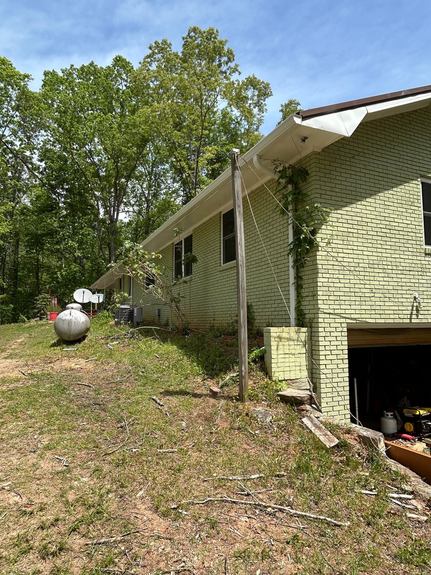 Side view of a light green house with a garage, trees, and a propane tank on a grassy slope.
