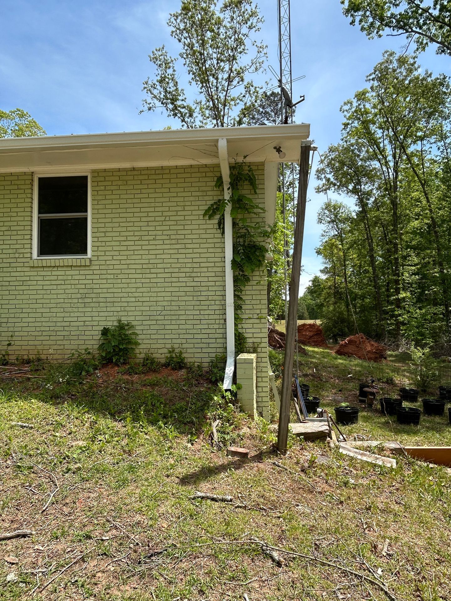 Side of a pale green house with a porch post, window, and small garden area under trees.