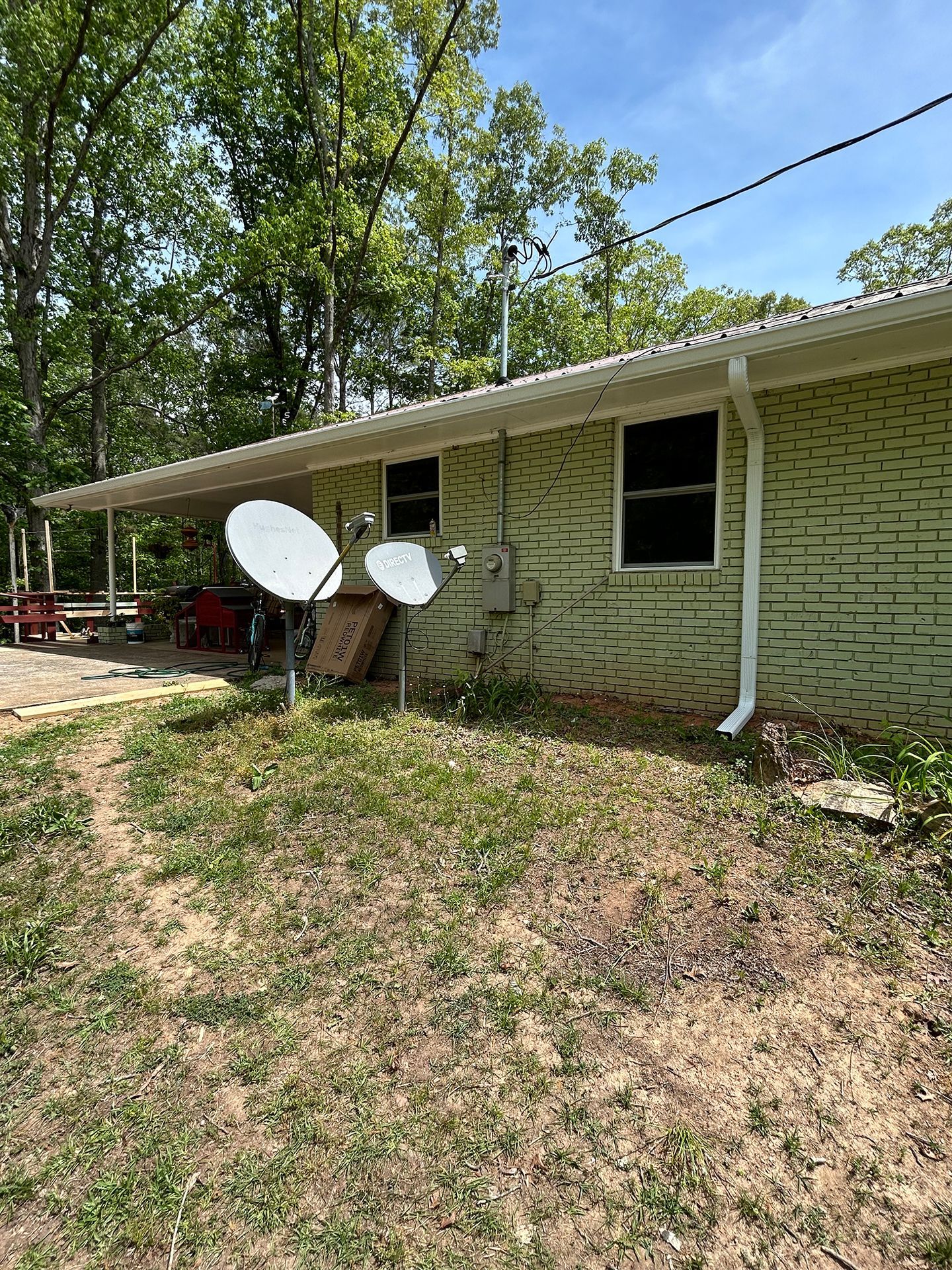 House side with two satellite dishes, window, downspout, and trees in the yard