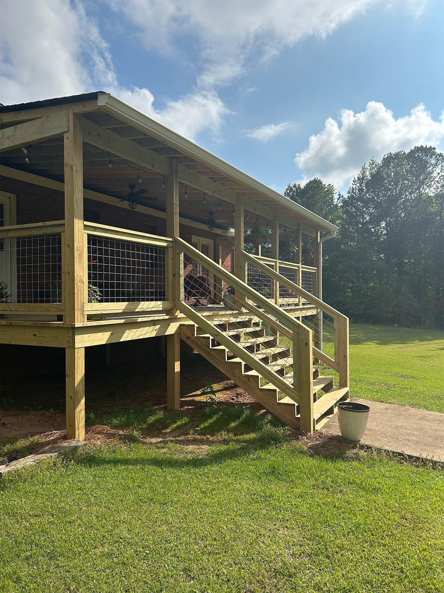 Wooden porch with stairs on a raised cabin, beside a grassy yard under a partly cloudy sky