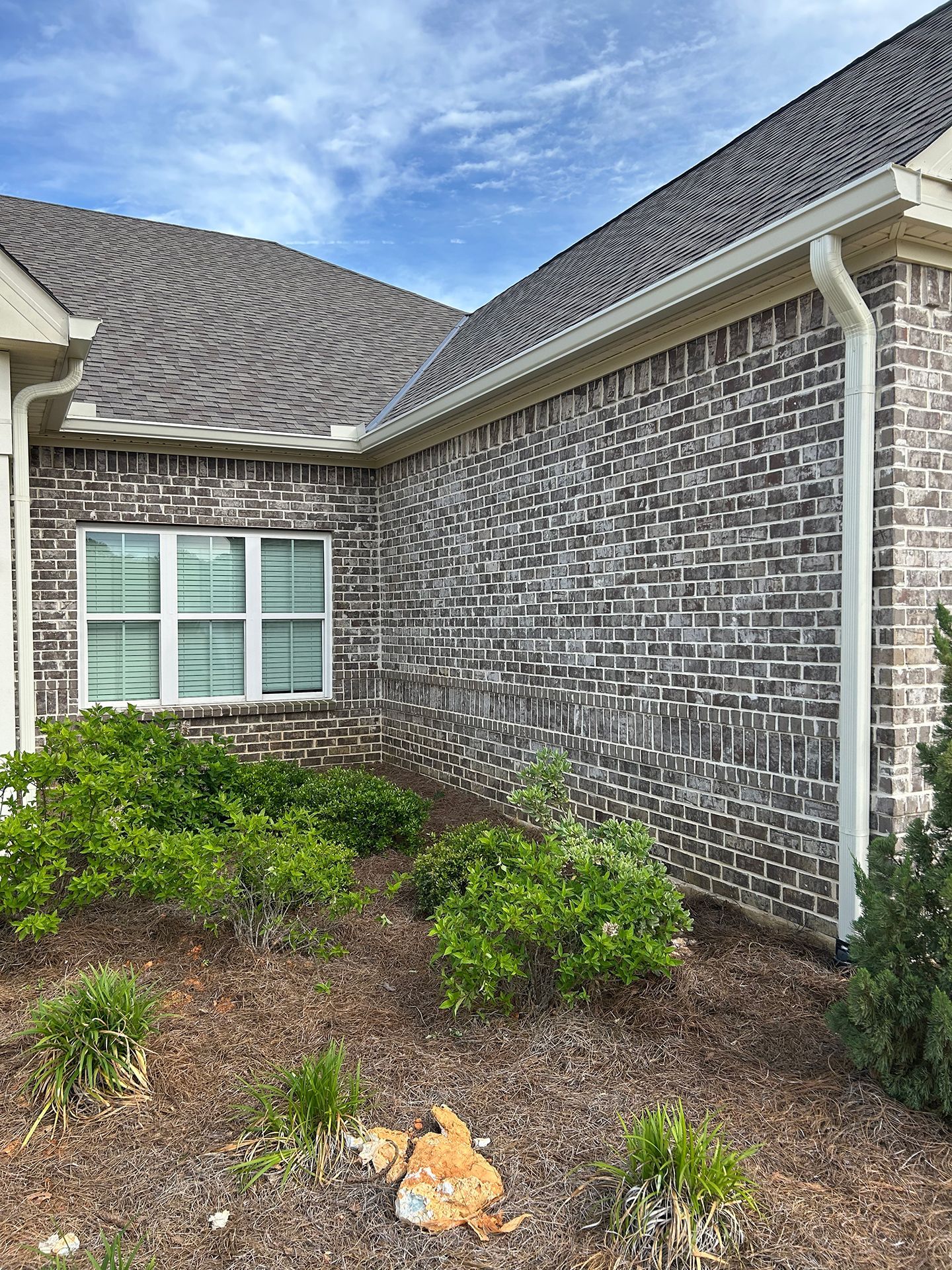 Brick house exterior with white-trimmed window, roofline, and landscaped shrubs under a blue sky