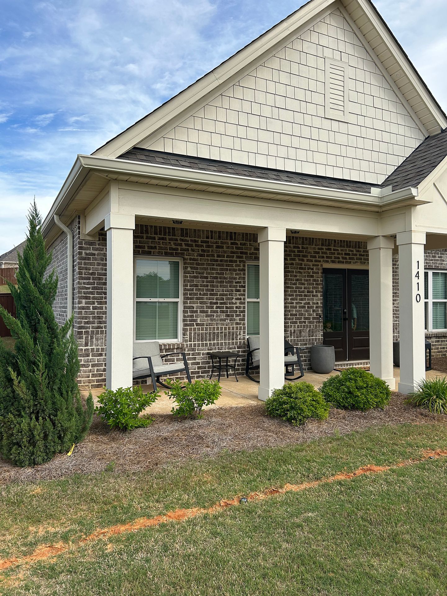 Front porch of a brick house with white columns, a gabled roof, and small shrubs along the lawn.
