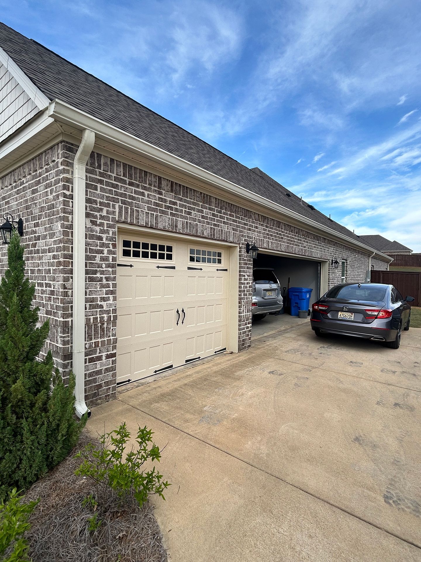 Brick house garage with beige doors, blue car parked inside, and a black sedan in the driveway.