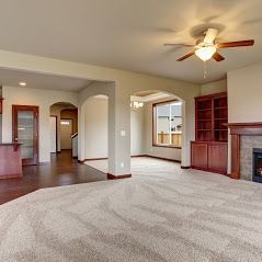 A spacious living room with beige carpeting, cream walls, an arched entryway, wooden bookshelves, and a ceiling fan.