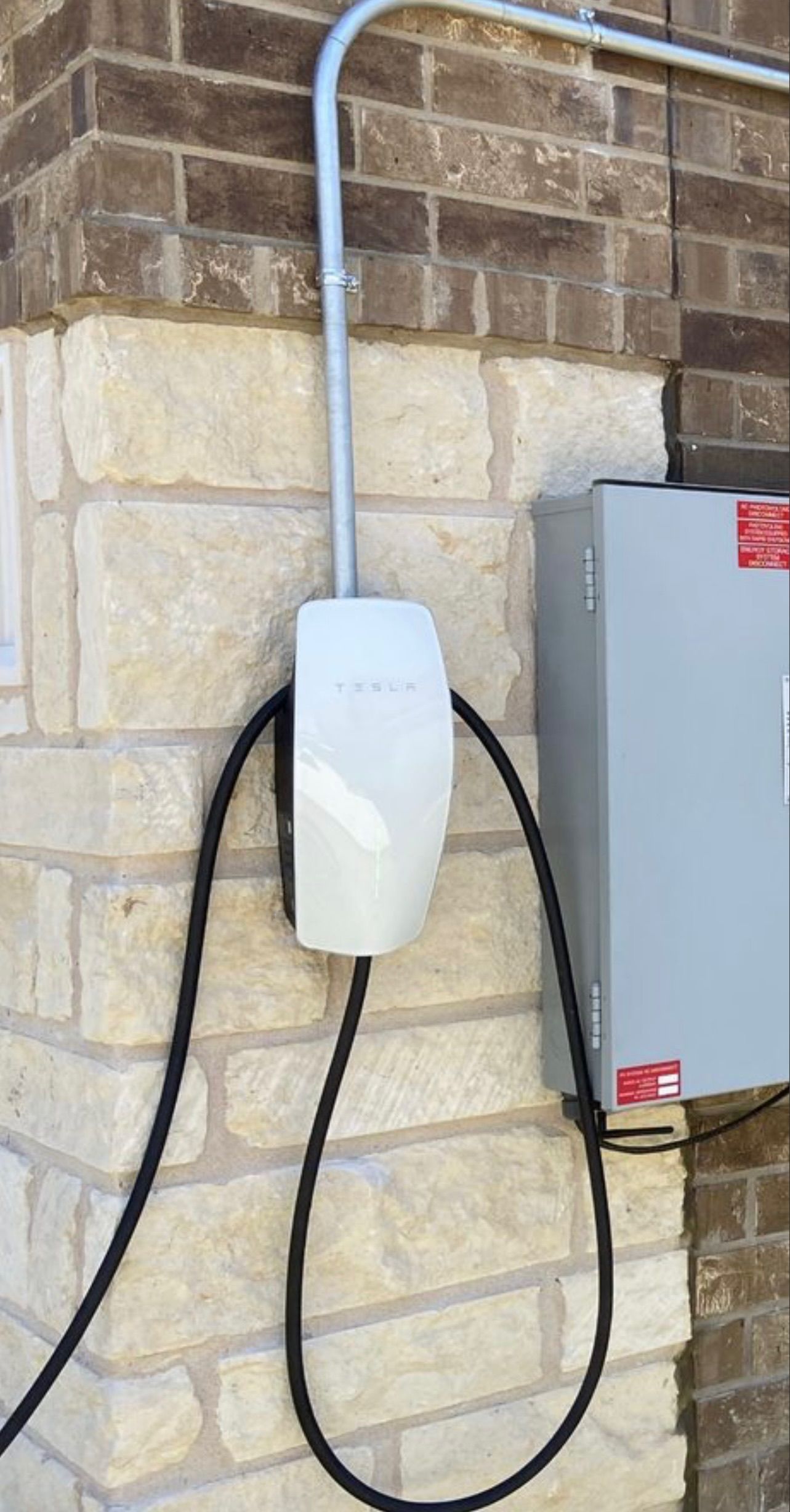 An electric vehicle charger mounted on a brick wall, connected to a gray conduit and a metal electrical box. 