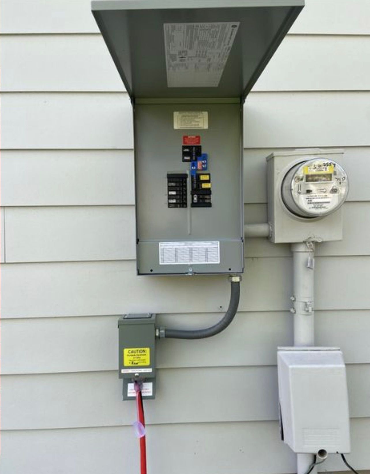 Electrical panel and meter box on a house exterior with the panel door open. A red cord plugs into a gray box below.