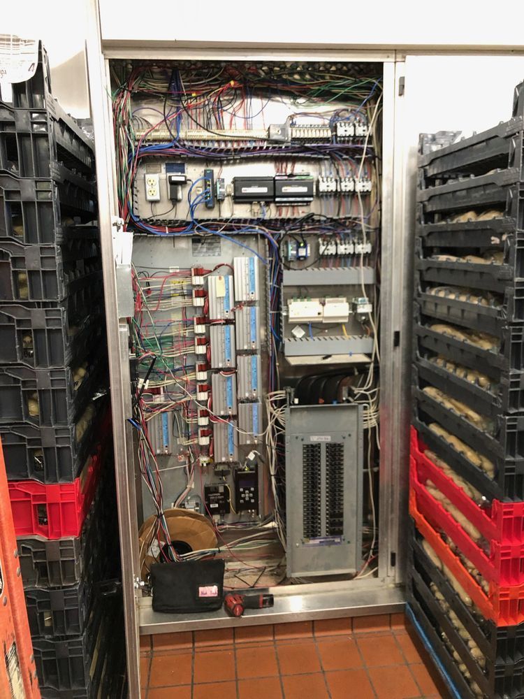 Electrical control panel with complex wiring inside a stainless steel cabinet, flanked by stacks of black plastic food trays.