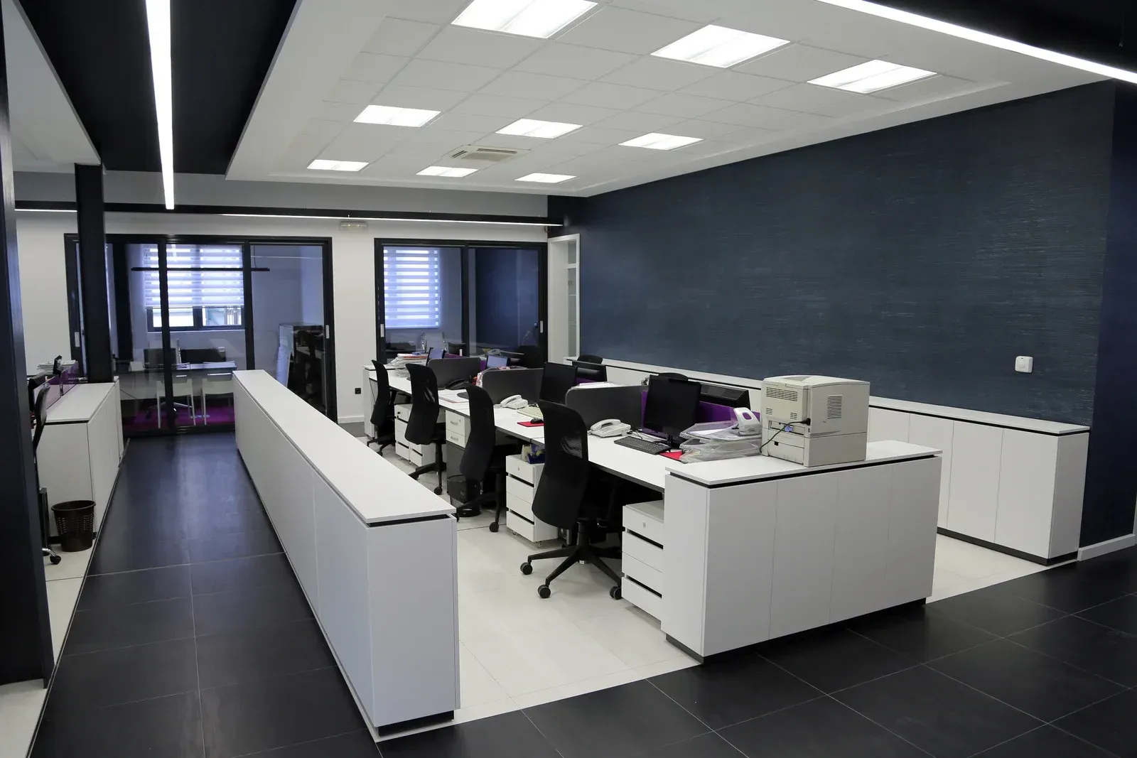 Modern office interior with multiple workstations, white desks, black chairs, and a navy blue accent wall.