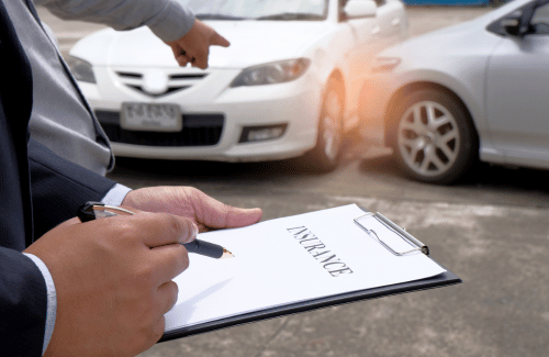 Person writing on a clipboard while inspecting a car accident scene with damaged white cars and another person pointing.