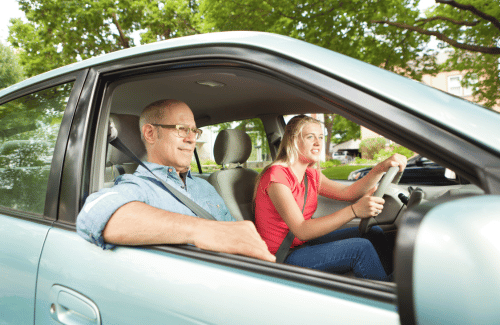 Two people inside a light blue car, smiling while driving along a tree-lined road.