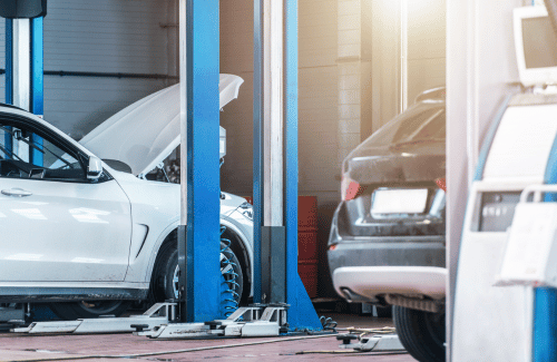 Cars in an auto repair garage, one white car with hood open beside a lift.