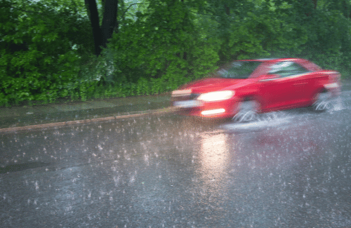 Red car driving through heavy rain on a wet road near green trees.