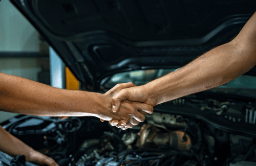Two people shaking hands in front of an open car hood in a repair shop