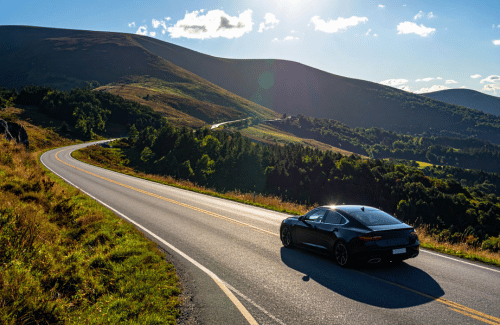 Black sedan driving along a winding mountain road through rolling green hills under bright sunshine.