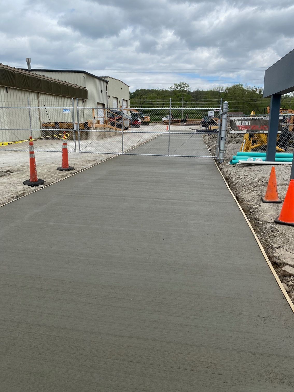 Freshly paved concrete path at a construction site, bordered by orange cones and equipment under cloudy skies