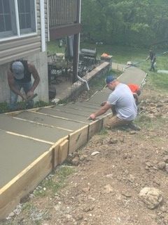 Workers Working On A Construction Site — St. Charles, MO — TC Concrete Services Workers pouring and smoothing a concrete walkway beside a house, with boards and a wheelbarrow nearby.