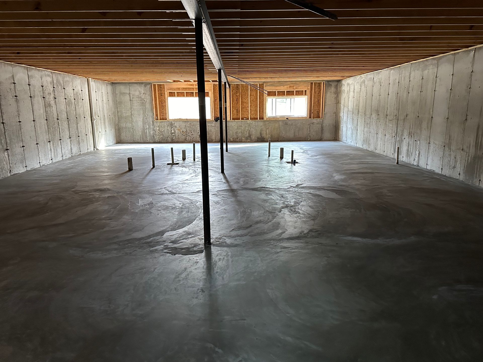 Empty unfinished basement with concrete floor, exposed wooden ceiling joists, and central support column.